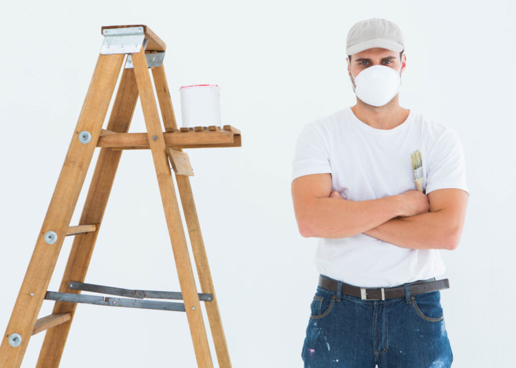 Portrait of man with paintbrush standing arms crossed by ladder on white background