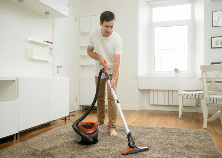 Happy smiling man vacuum cleaning the carpet in the living room, modern scandinavian interior. Busy, cleaning day. Home, housekeeping concept.