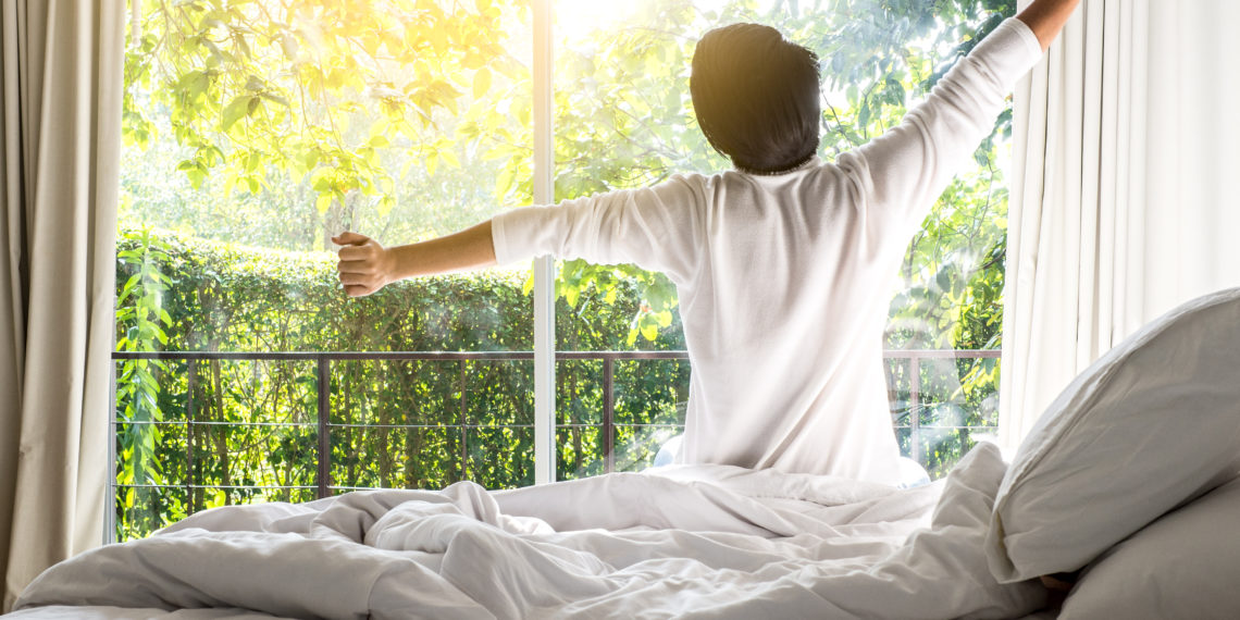 man happy waking up in the bed rising hands to window in the morning with fresh feeling relax