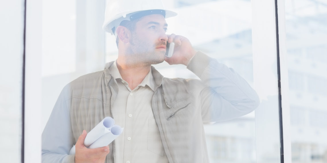 Male architect with blueprints using mobile phone in office