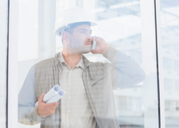 Male architect with blueprints using mobile phone in office