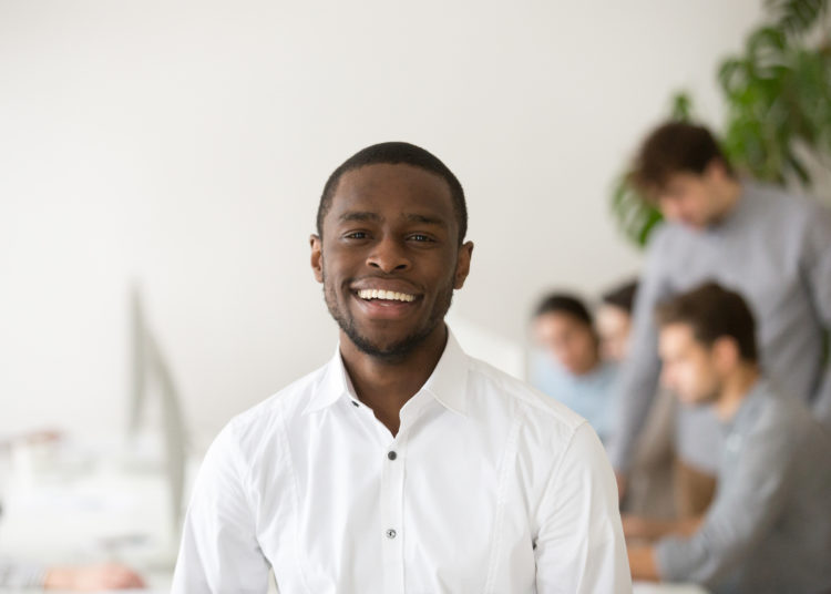 Happy african american professional smiling looking at camera with colleagues at background, friendly black business coach, successful intern, team leader member posing in office, head shot portrait