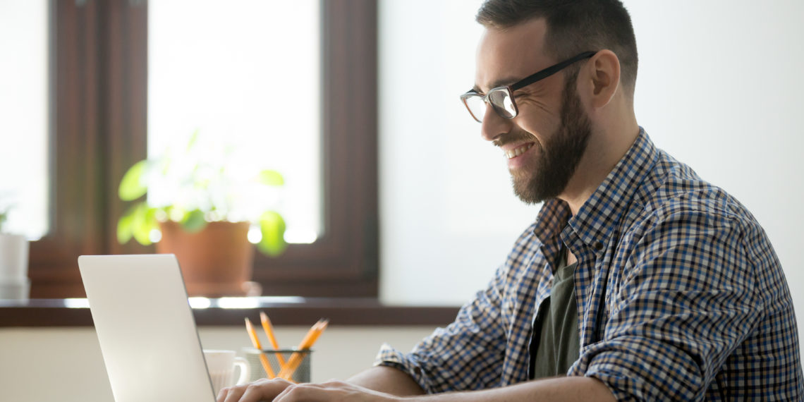 Happy male office worker smiling typing message at laptop, chatting with friends, writing positive email or consulting client online. Concept of laugher, positive work atmosphere, help and assistance