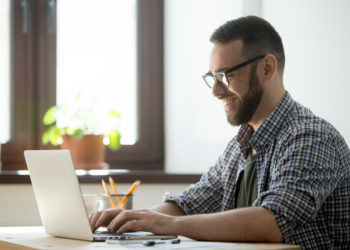 Happy male office worker smiling typing message at laptop, chatting with friends, writing positive email or consulting client online. Concept of laugher, positive work atmosphere, help and assistance