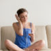 Teen girl wearing headphones enjoying new popular mp3 music on mobile phone application sitting on sofa at home, young smiling woman listening to favorite audio tracks playlist on smartphone player