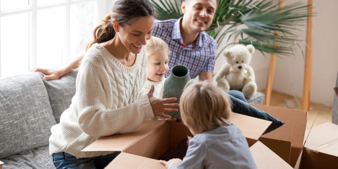 Happy family with two kids unpacking boxes after relocation moving into or settling in new home concept, excited small children helping parents with belongings sitting on sofa in living room together