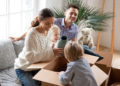 Happy family with two kids unpacking boxes after relocation moving into or settling in new home concept, excited small children helping parents with belongings sitting on sofa in living room together