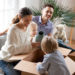 Happy family with two kids unpacking boxes after relocation moving into or settling in new home concept, excited small children helping parents with belongings sitting on sofa in living room together
